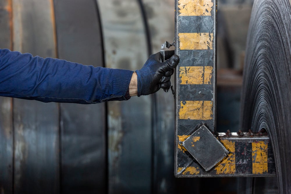 Worker using a measuring tool on industrial metal coils, emphasizing precision in rigging and lifting operations related to shackles and chains.