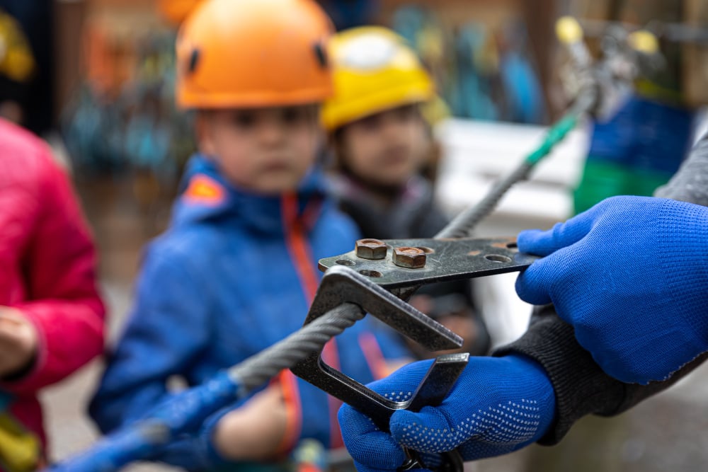 Person using rigging equipment to demonstrate safety practices, with children in helmets observing, emphasizing maintenance and inspection of rigging gear.